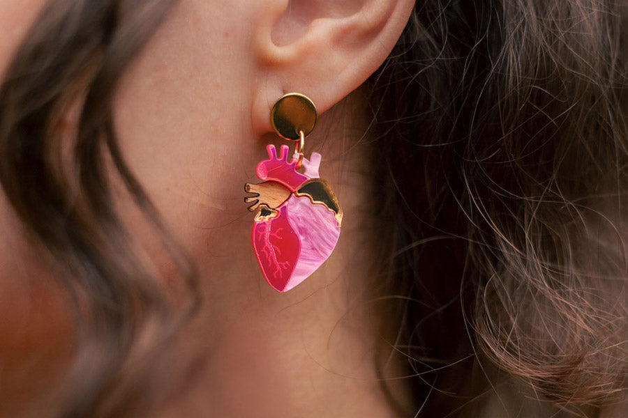 A close-up of a persons ear wearing a colorful anatomical heart-shaped earring with pink, gold, and red details. Curly brown hair frames the ear.