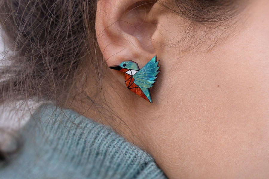 A close-up of a person wearing a colorful geometric bird-shaped earring, featuring shades of blue, green, and orange, against their ear. The person is wearing a light blue sweater.