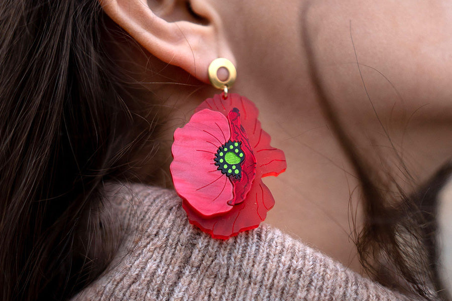 Close-up of a persons ear wearing a large, red, flower-shaped earring with green and black details, paired with a beige knit sweater.
