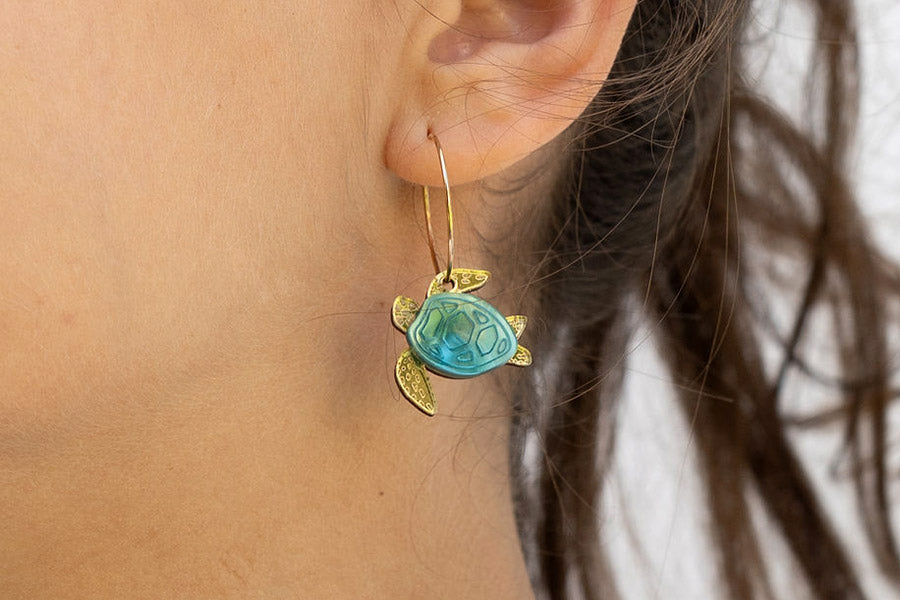 A close-up of a person wearing a gold and blue turtle-shaped earring. The earring features gold flippers and head with a turquoise blue shell, hanging from a simple wire hook.