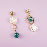A pair of mismatched earrings on a light purple background; both feature white floral designs, gold accents, and green beads—blending modern charm with timeless elegance in their circular and oval shapes.