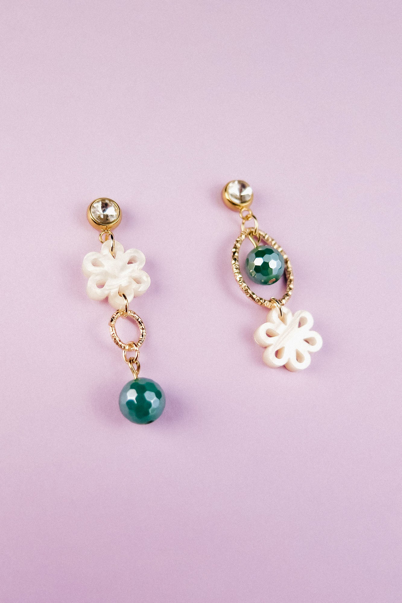 A pair of mismatched earrings on a light purple background; both feature white floral designs, gold accents, and green beads—blending modern charm with timeless elegance in their circular and oval shapes.