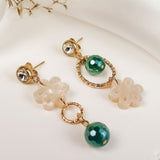 A pair of mismatched gold earrings with green beads and cream flower-shaped charms exudes timeless elegance, displayed on a white surface with dried grass in the background.