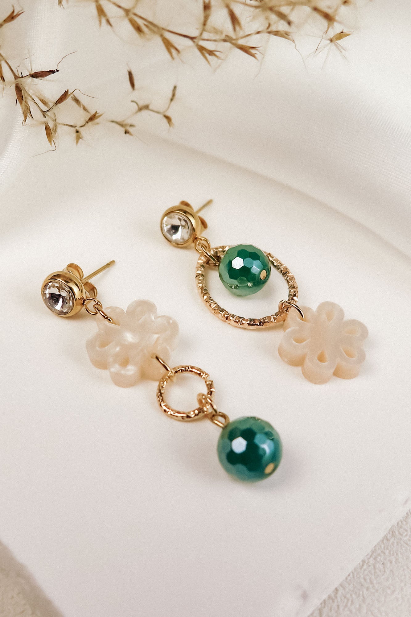 A pair of mismatched gold earrings with green beads and cream flower-shaped charms exudes timeless elegance, displayed on a white surface with dried grass in the background.