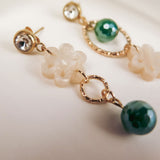 Close-up of two mismatched earrings featuring a clear gemstone stud, a gold ring, a white flower-shaped bead, and a green faceted bead, all laid out on a soft, neutral background for modern charm.
