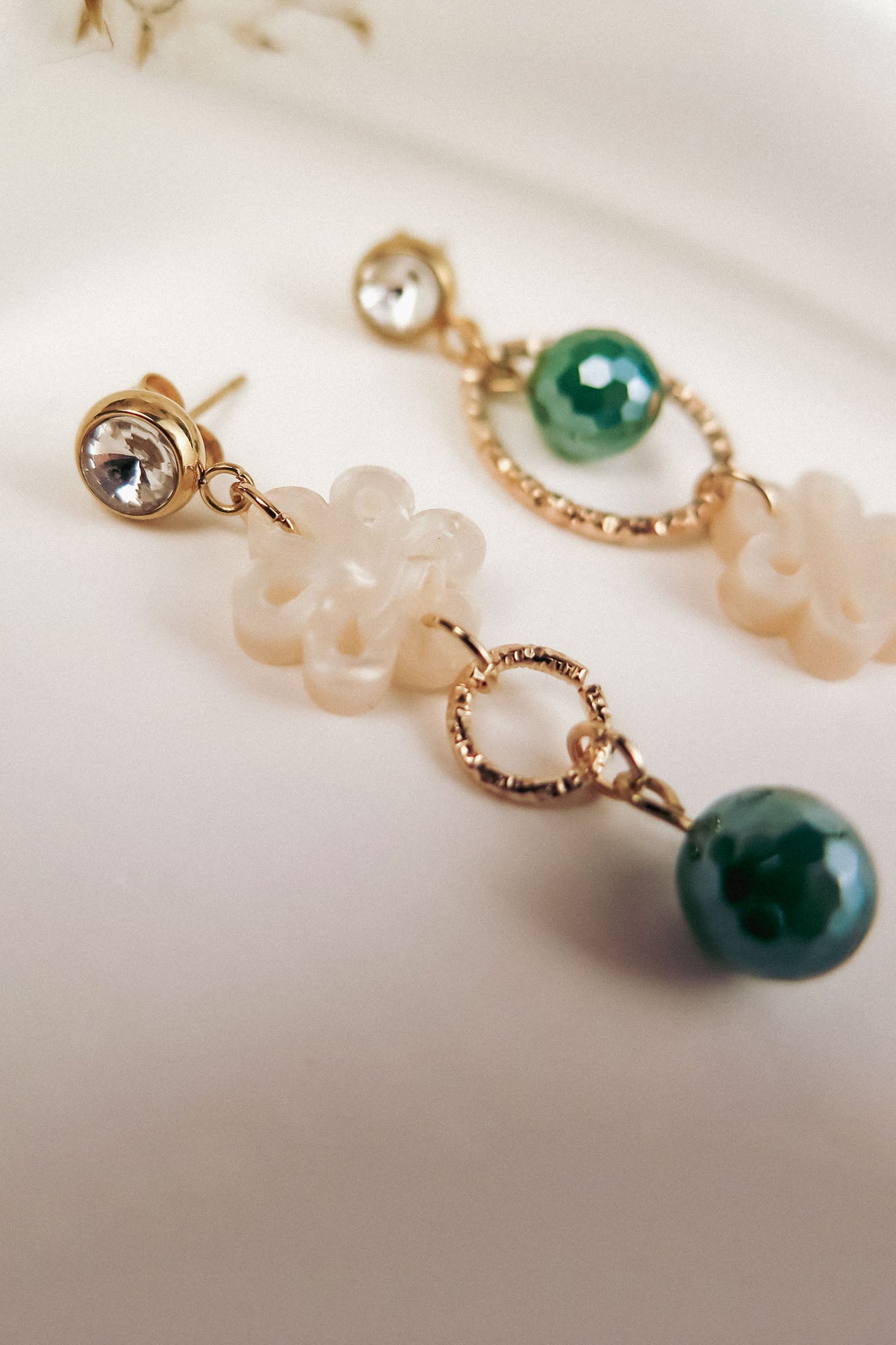 Close-up of two mismatched earrings featuring a clear gemstone stud, a gold ring, a white flower-shaped bead, and a green faceted bead, all laid out on a soft, neutral background for modern charm.