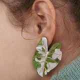 A close-up of a persons ear adorned with large, leaf-shaped Alocasia Earrings in green and white. The hand-crafted jewelry stands out against their brown hair and a light blue-green garment.