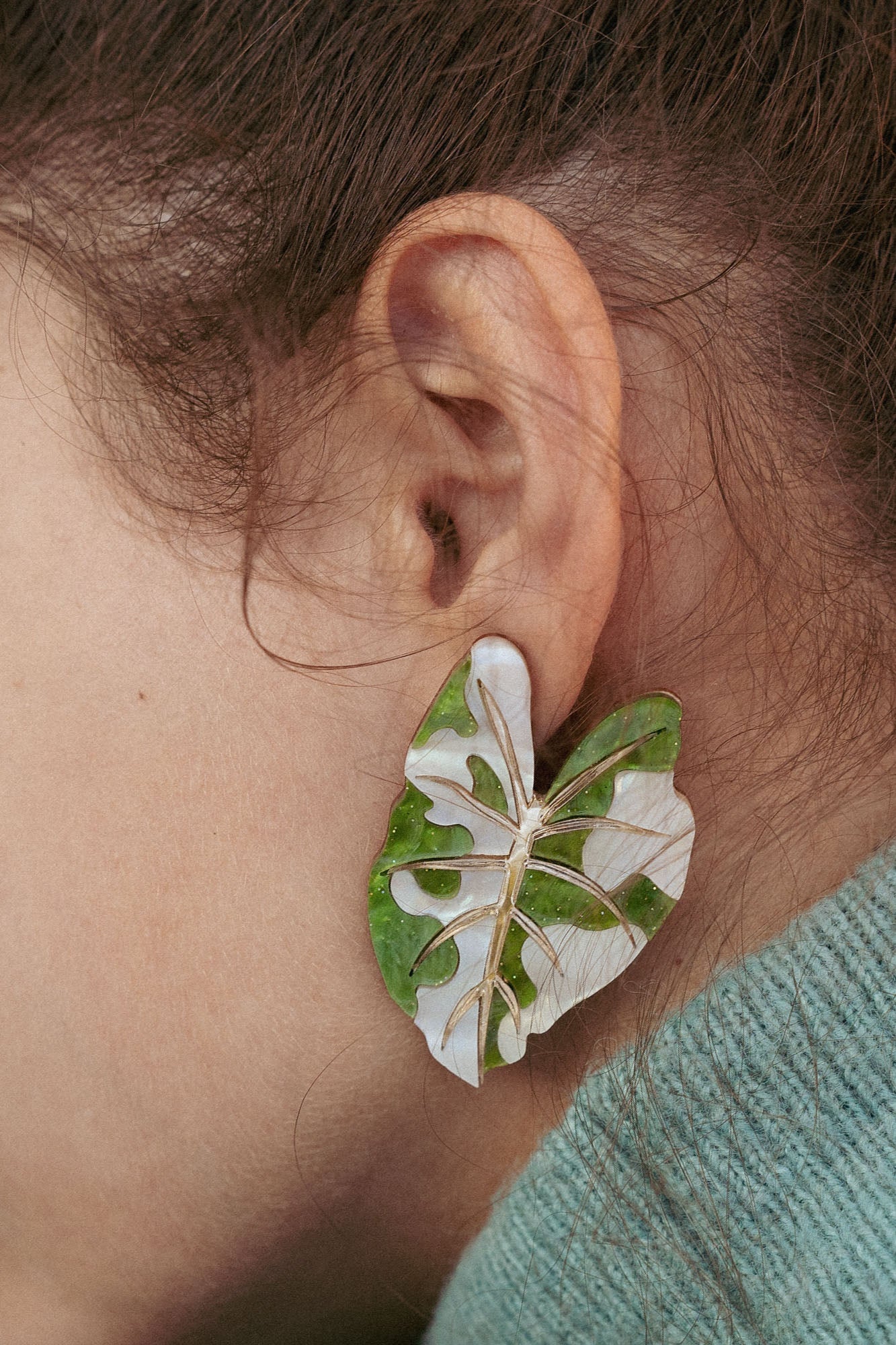 A close-up of a persons ear adorned with large, leaf-shaped Alocasia Earrings in green and white. The hand-crafted jewelry stands out against their brown hair and a light blue-green garment.