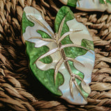 A close-up of Alocasia Earrings, shaped like a green and white leaf with metallic veins, resting on a woven, textured surface—perfect for those who love hand-crafted jewelry.