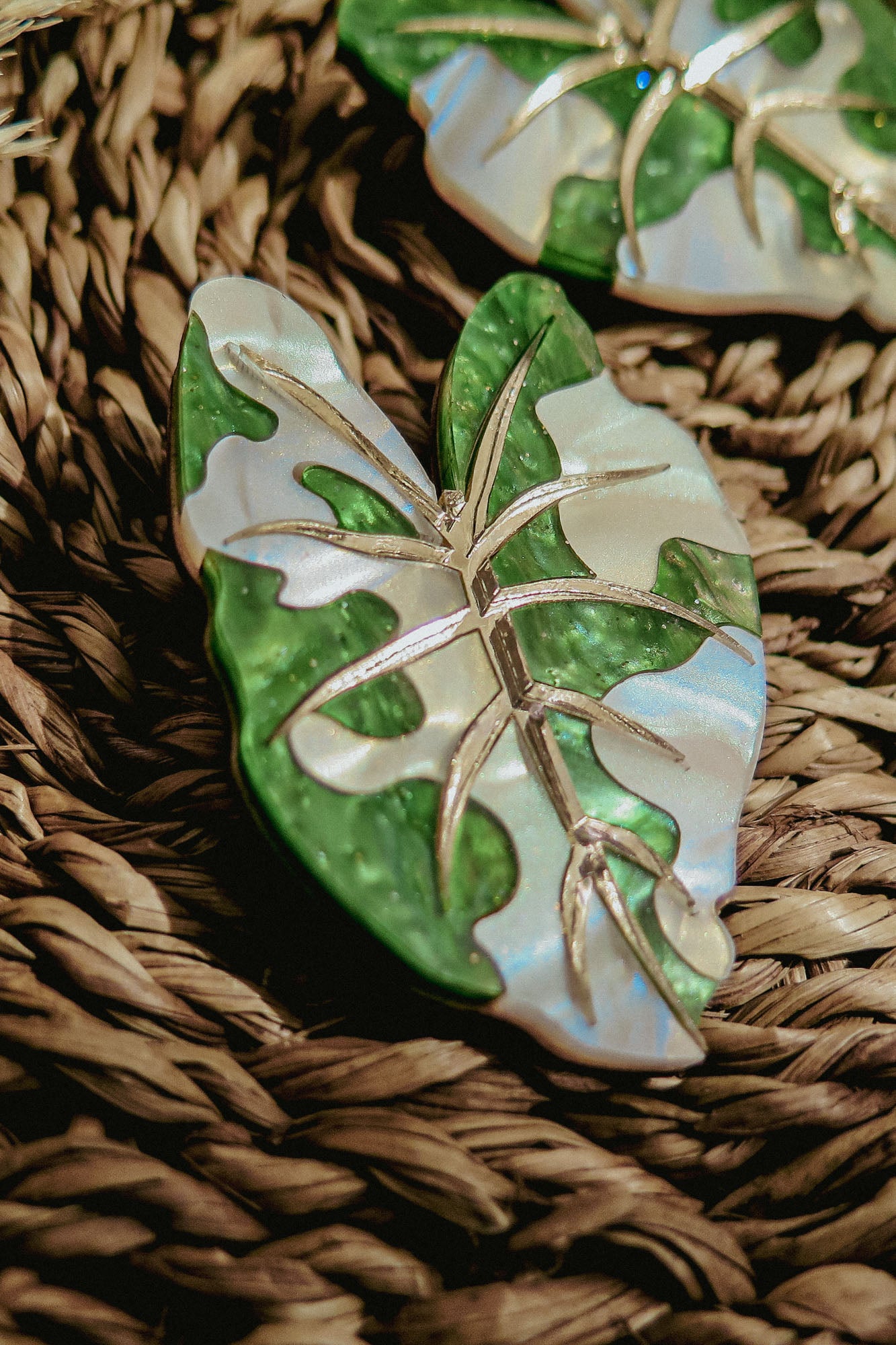 A close-up of Alocasia Earrings, shaped like a green and white leaf with metallic veins, resting on a woven, textured surface—perfect for those who love hand-crafted jewelry.