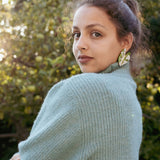 A woman with curly hair, wearing a light blue knit sweater and large Alocasia Earrings, stands outdoors, looking over her shoulder. Sunlight filters through green trees in the background, highlighting her stylish hand-crafted jewelry.