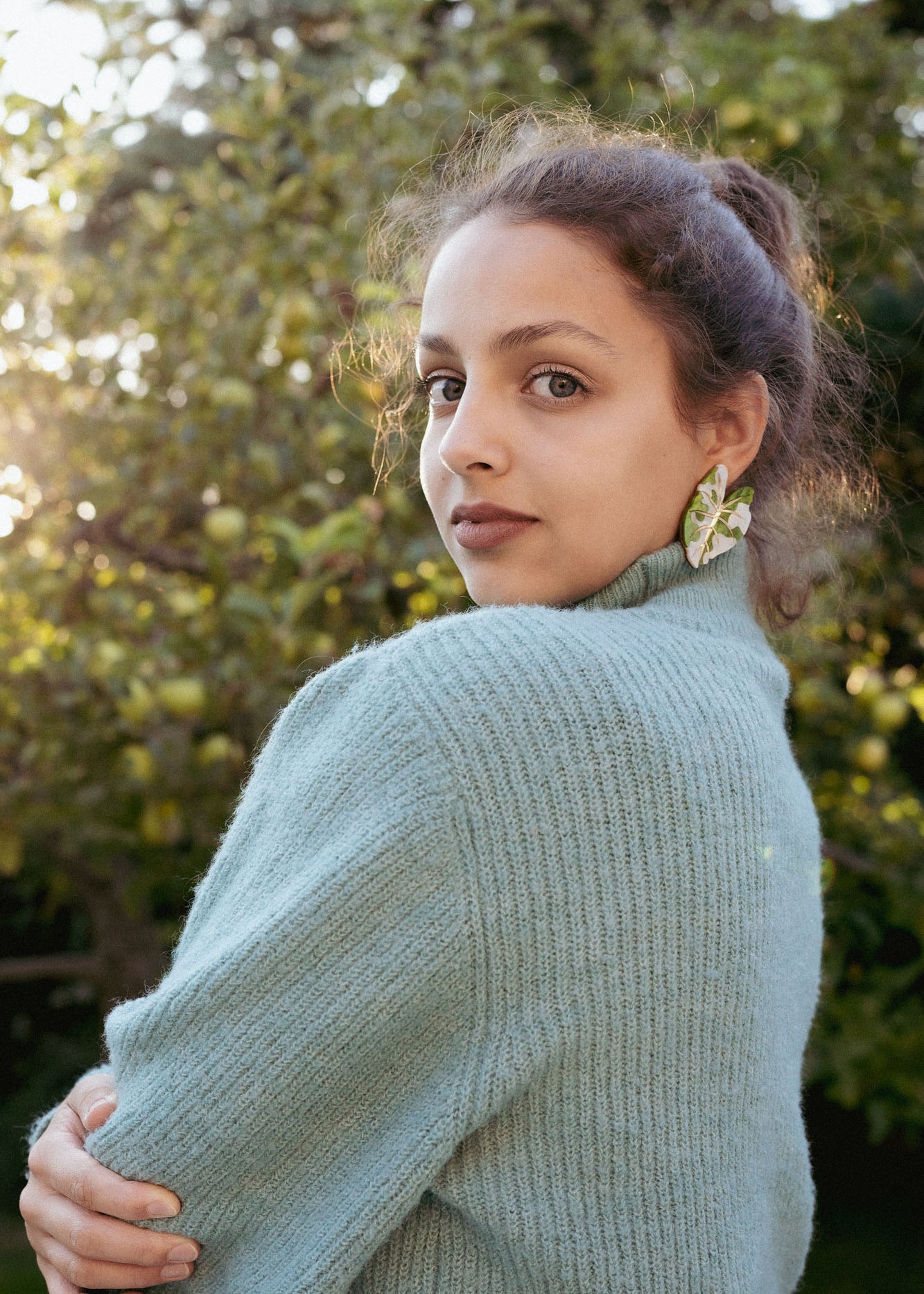 A woman with curly hair, wearing a light blue knit sweater and large Alocasia Earrings, stands outdoors, looking over her shoulder. Sunlight filters through green trees in the background, highlighting her stylish hand-crafted jewelry.