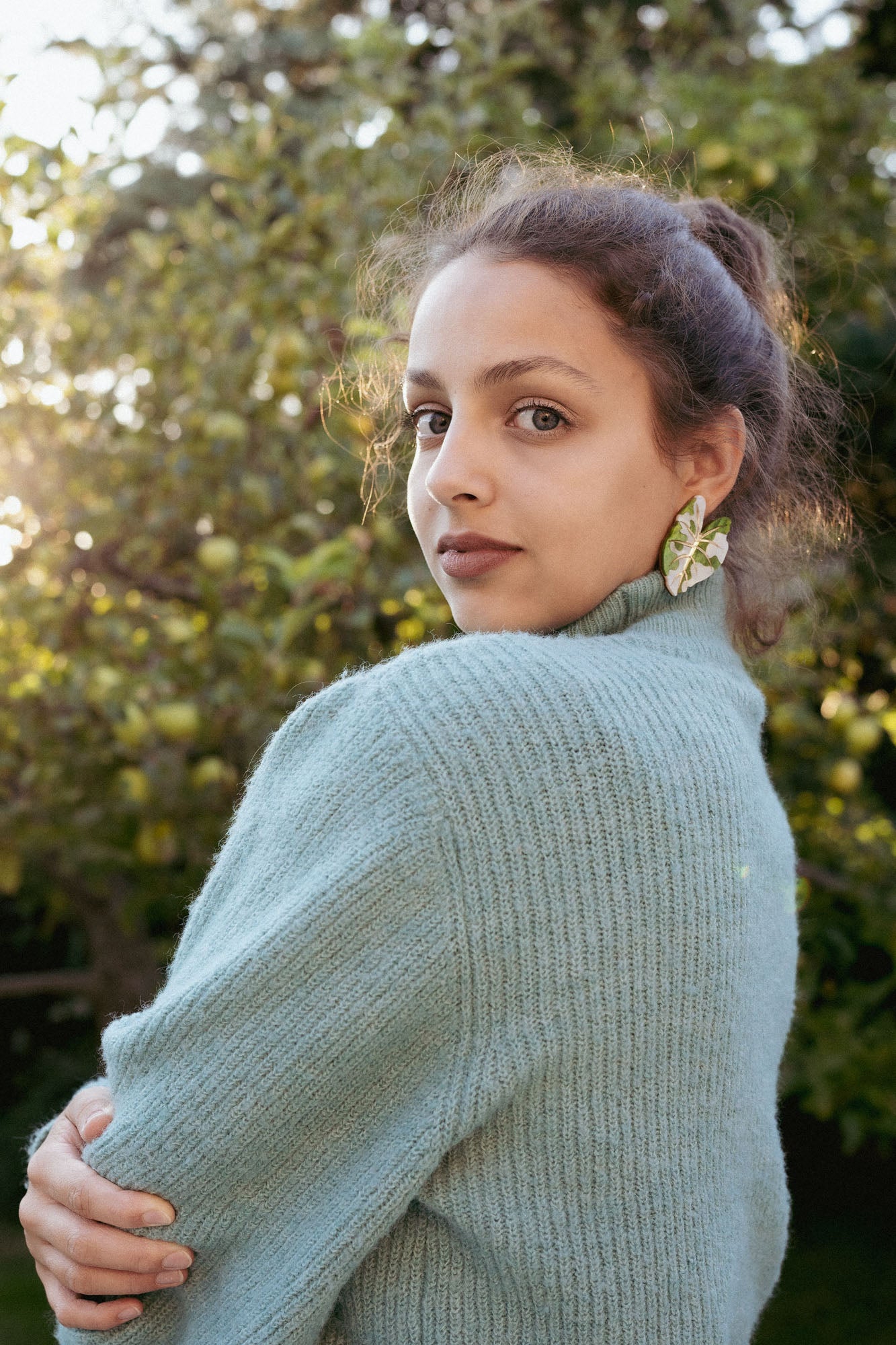 A woman with curly hair, wearing a light blue knit sweater and large Alocasia Earrings, stands outdoors, looking over her shoulder. Sunlight filters through green trees in the background, highlighting her stylish hand-crafted jewelry.