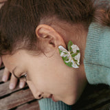 A person with brown hair lies on a wooden surface, wearing a green sweater. An Alocasia Earrings-inspired green and white leaf, reminiscent of hand-crafted jewelry, is placed over their ear, resembling an earring.