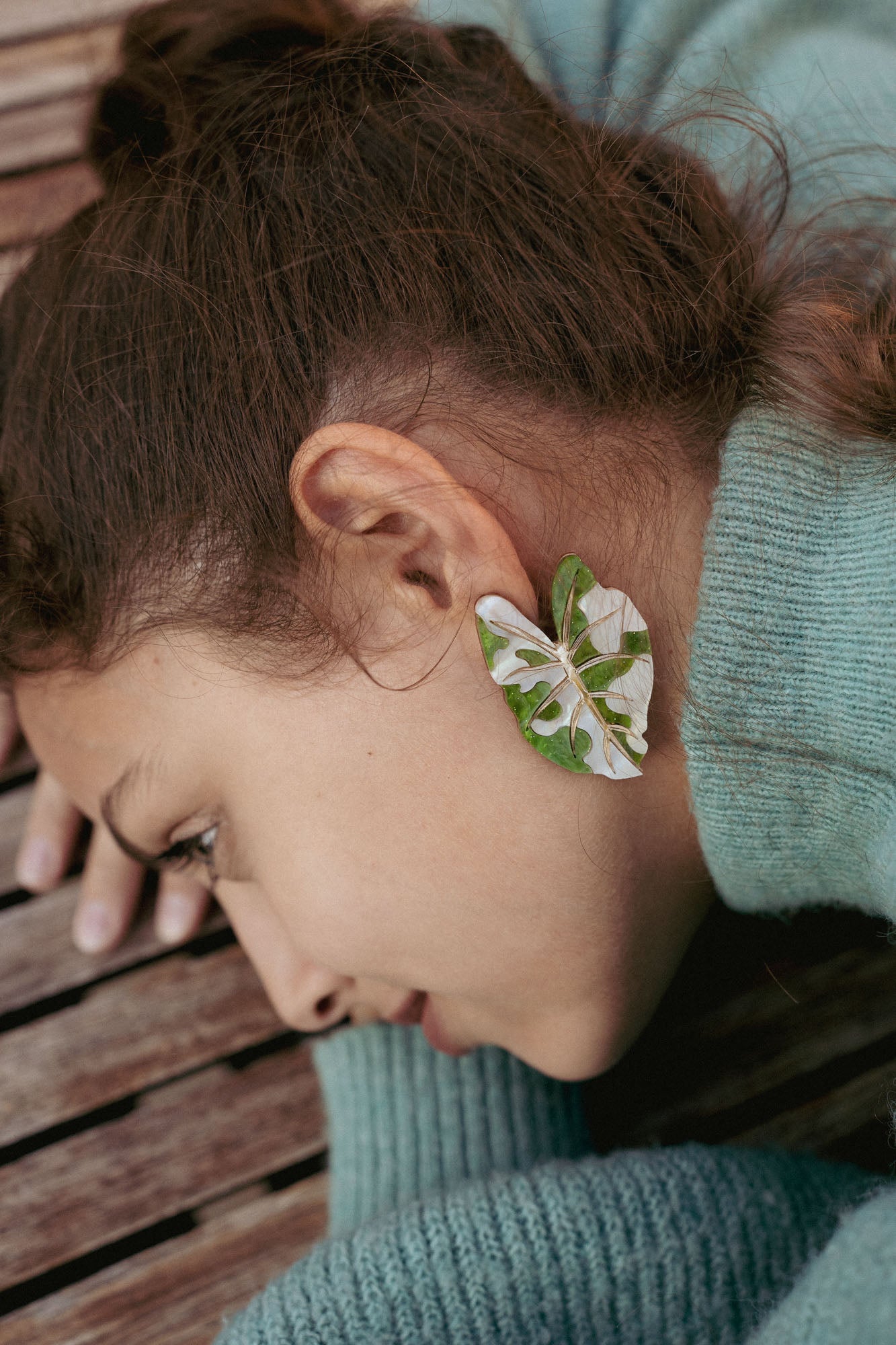 A person with brown hair lies on a wooden surface, wearing a green sweater. An Alocasia Earrings-inspired green and white leaf, reminiscent of hand-crafted jewelry, is placed over their ear, resembling an earring.
