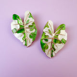 Two decorative pins shaped like monstera leaves, inspired by hand-crafted jewelry, feature a mix of green and white colors with gold vein detailing, and are placed on a light purple background.
