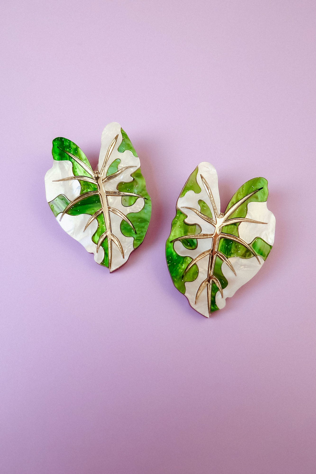 Two decorative pins shaped like monstera leaves, inspired by hand-crafted jewelry, feature a mix of green and white colors with gold vein detailing, and are placed on a light purple background.