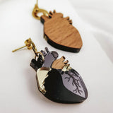 A close-up of two Anatomical Heart Earrings, handcrafted from wood and black acrylic with gold hooks, showcases unique handcrafted accessories and a hint of gothic flair on a white surface.
