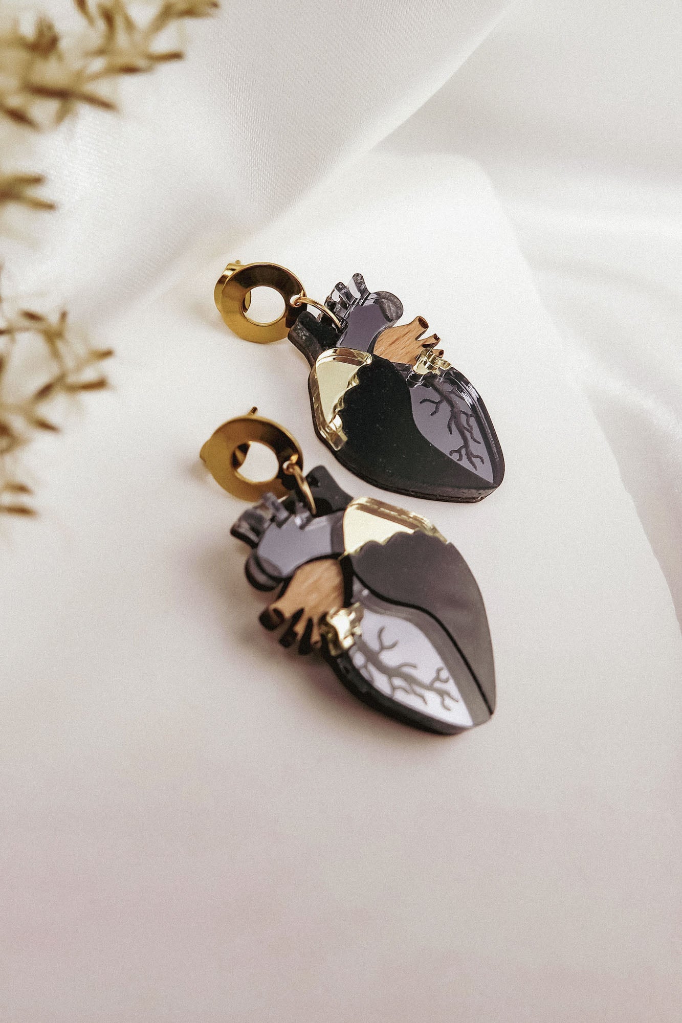 Two anatomical heart earrings with black, silver, and gold accents add a touch of gothic flair, lying on white fabric beside delicate dried flowers—unique handcrafted accessories for those who love bold style.