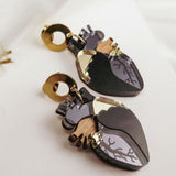 A pair of Anatomical Heart Earrings with gold accents and black, grey, and wood-toned details adds a touch of gothic flair, displayed on a white fabric background—perfect for lovers of unique handcrafted accessories.
