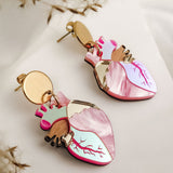 Anatomical Heart Earrings in a bold style, shaped with pink, red, and metallic details, lie on a soft white surface surrounded by dried plant stems.