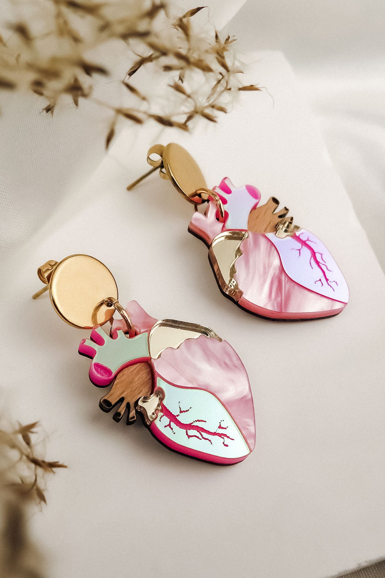 Anatomical Heart Earrings in a bold style, shaped with pink, red, and metallic details, lie on a soft white surface surrounded by dried plant stems.