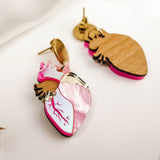 Anatomical Heart Earrings crafted from wood and pink marbled acrylic, accented with gold-tone metal in a bold style, are displayed on a white fabric background.