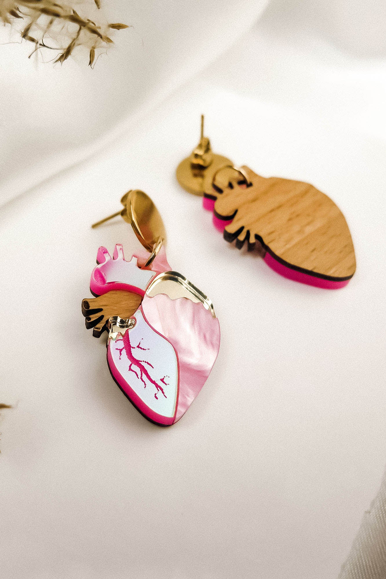 Anatomical Heart Earrings crafted from wood and pink marbled acrylic, accented with gold-tone metal in a bold style, are displayed on a white fabric background.
