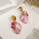A pair of Anatomical Heart Earrings in a bold style, shaped like hearts with pink and gold details, are displayed on a white fabric surface with dried grass in the background.