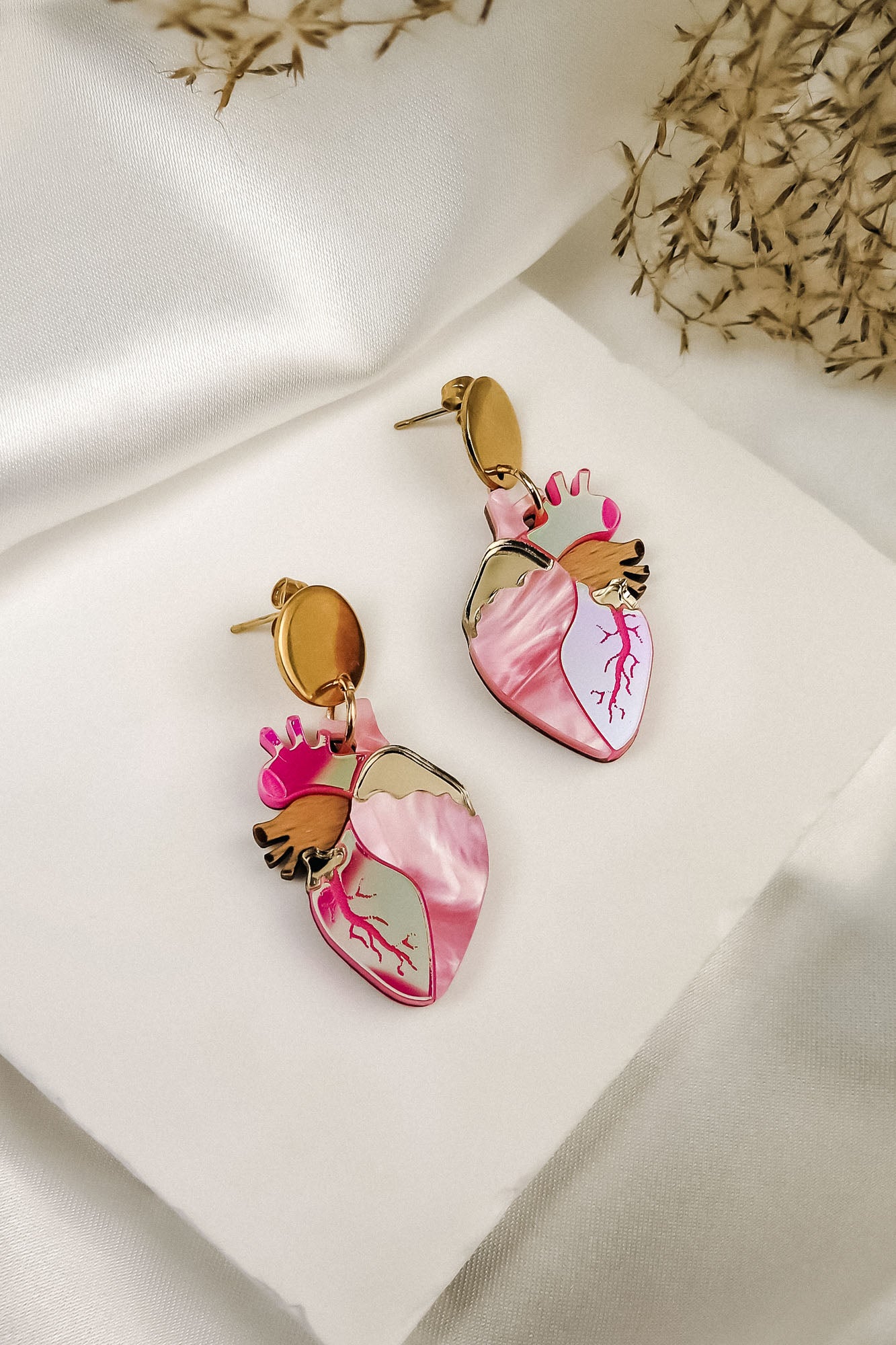 A pair of Anatomical Heart Earrings in a bold style, shaped like hearts with pink and gold details, are displayed on a white fabric surface with dried grass in the background.