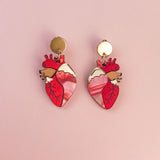 A pair of gold and pink Anatomical Heart Earrings, crafted in acrylic with detailed vein patterns and a unique style, displayed on a light pink background.