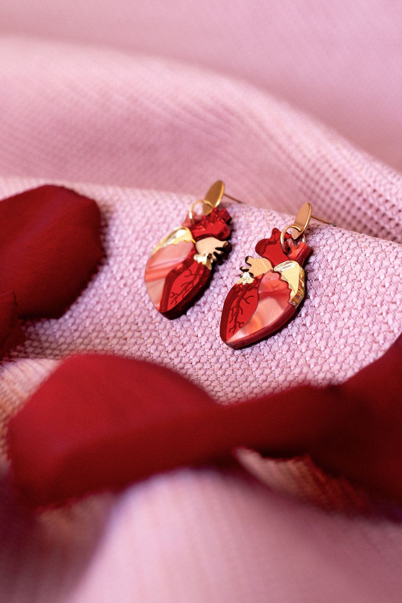 Two red Anatomical Heart Earrings with gold accents rest on a soft, pink textured fabric, surrounded by red rose petals—a unique style statement for acrylic earrings lovers.
