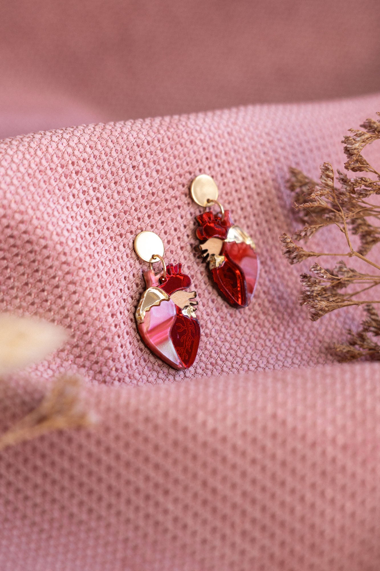 A pair of red and gold Anatomical Heart Earrings rest on textured pink fabric, with dried flowers nearby—acrylic earrings perfect for expressing your unique style.