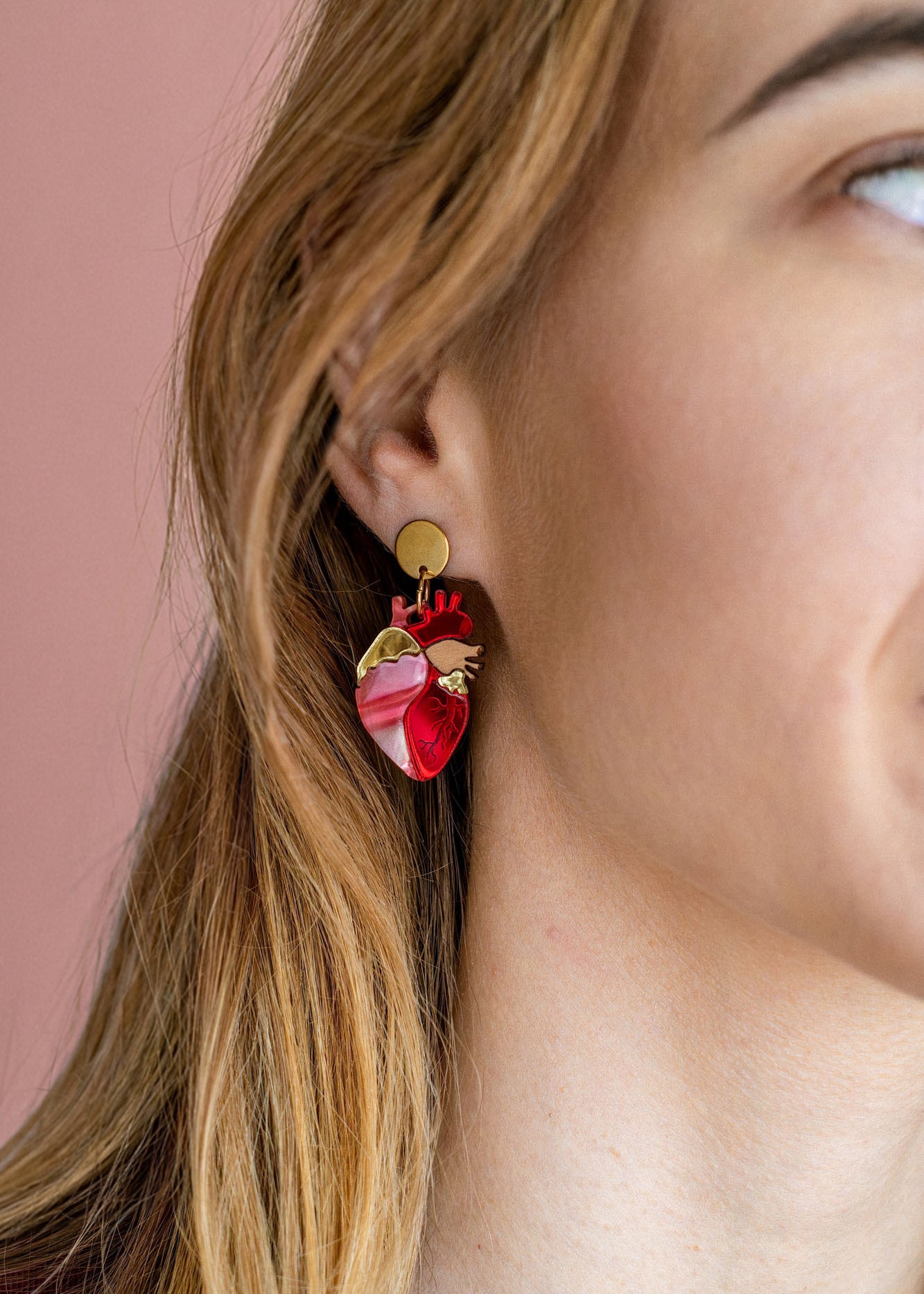 Close-up of a woman’s ear wearing unique style Anatomical Heart Earrings in red and pink tones with gold accents; her light brown hair is tucked behind her ear against a pink background, highlighting these striking acrylic earrings.