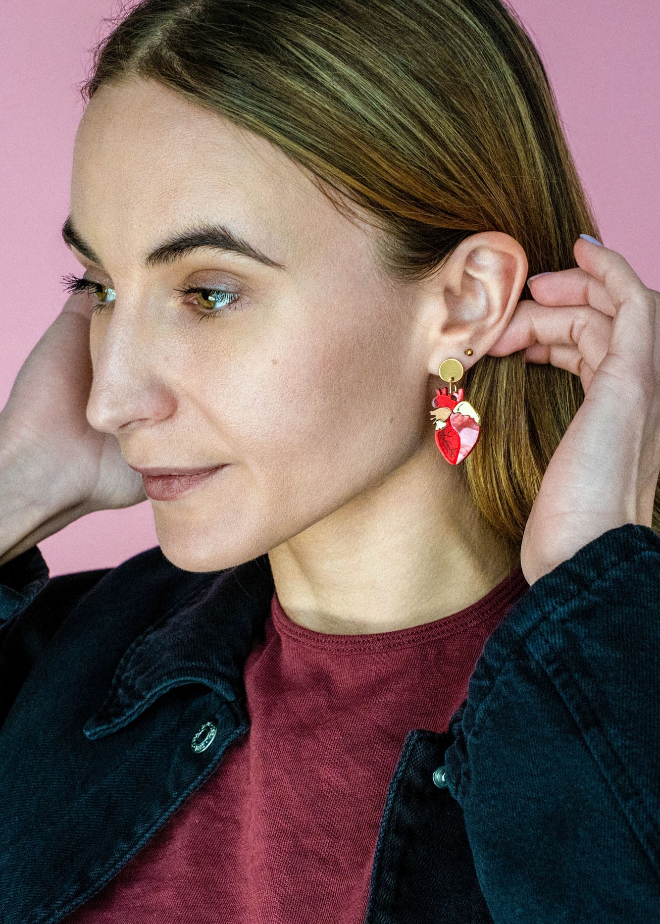 A woman with light brown hair wears a maroon shirt and black jacket, adjusting her unique acrylic earrings. She stands in front of a pink background, looking to the side with a slight smile.