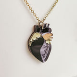 A gold chain necklace with a pendant shaped like an anatomical heart, featuring black, gold, and wood-toned sections etched with vein-like details. This handcrafted jewelry piece rests against a white background.