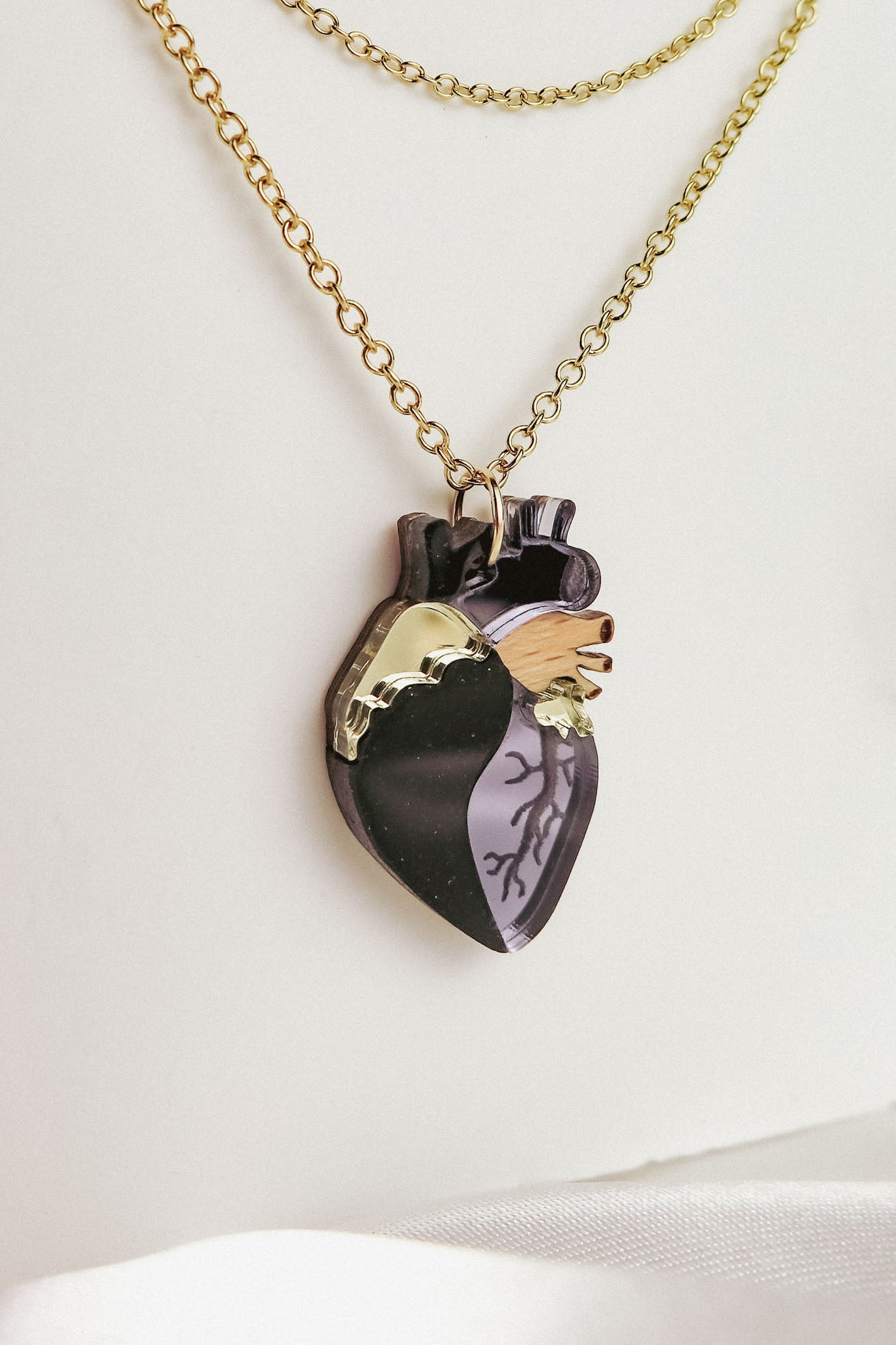 A gold chain necklace with a pendant shaped like an anatomical heart, featuring black, gold, and wood-toned sections etched with vein-like details. This handcrafted jewelry piece rests against a white background.