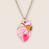 A gold chain anatomical heart necklace with a pink pendant featuring gold, orange, and dark pink details, displayed against a light beige background—perfect as a unique jewelry piece.