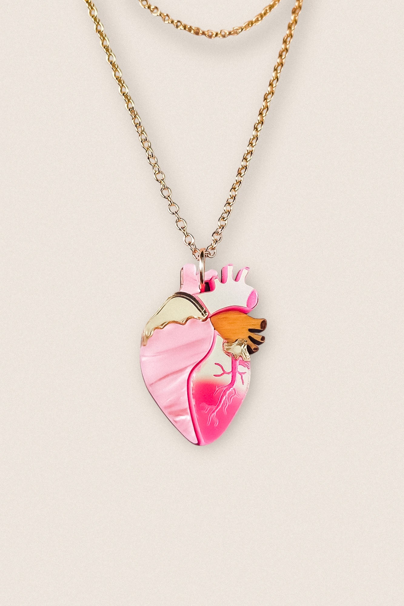 A gold chain anatomical heart necklace with a pink pendant featuring gold, orange, and dark pink details, displayed against a light beige background—perfect as a unique jewelry piece.