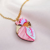 A unique jewelry piece, this gold chain necklace features a pendant shaped like an anatomical heart with pink, white, and gold details, beautifully displayed on soft, light-colored satin fabric.