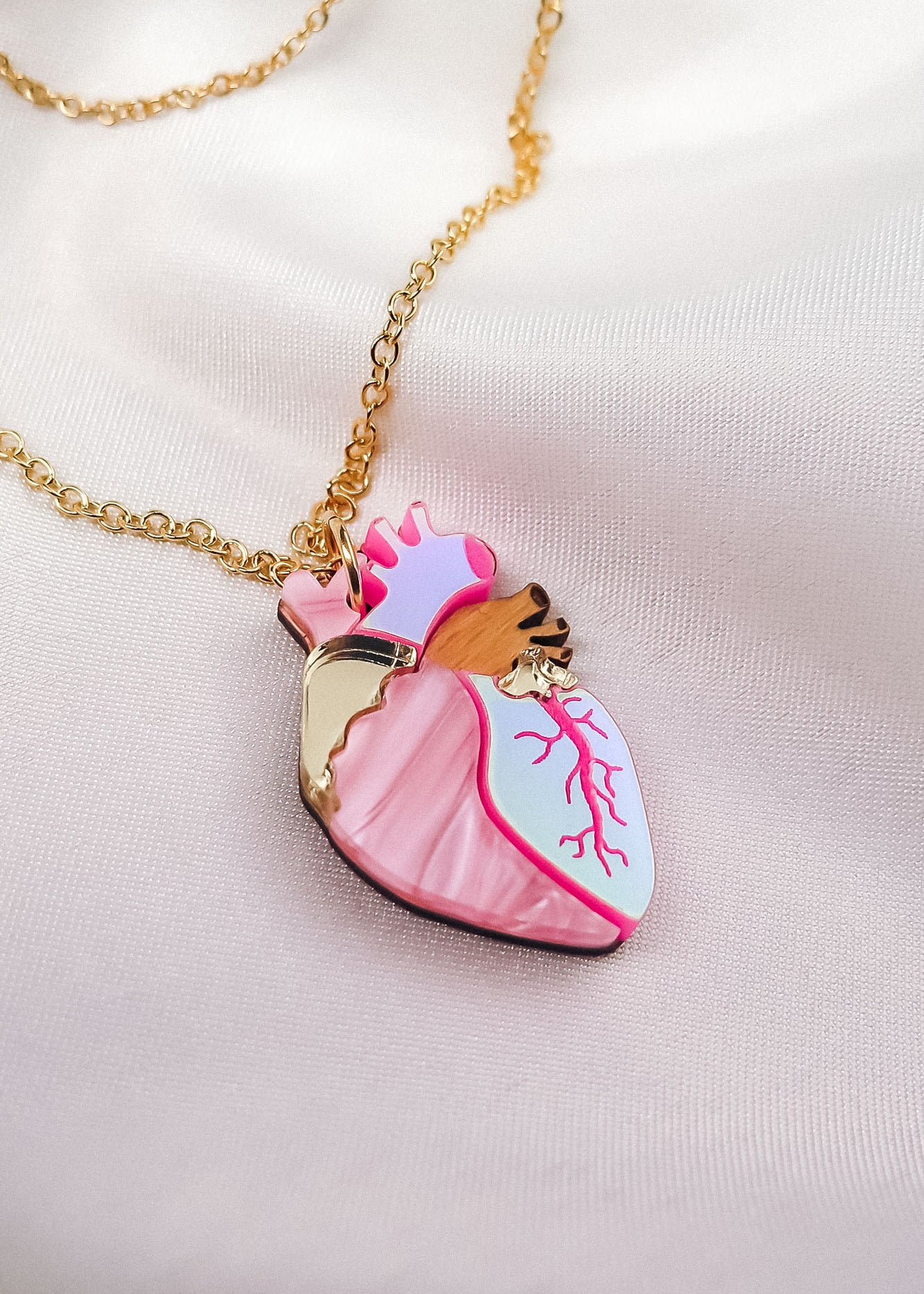 A unique jewelry piece, this gold chain necklace features a pendant shaped like an anatomical heart with pink, white, and gold details, beautifully displayed on soft, light-colored satin fabric.