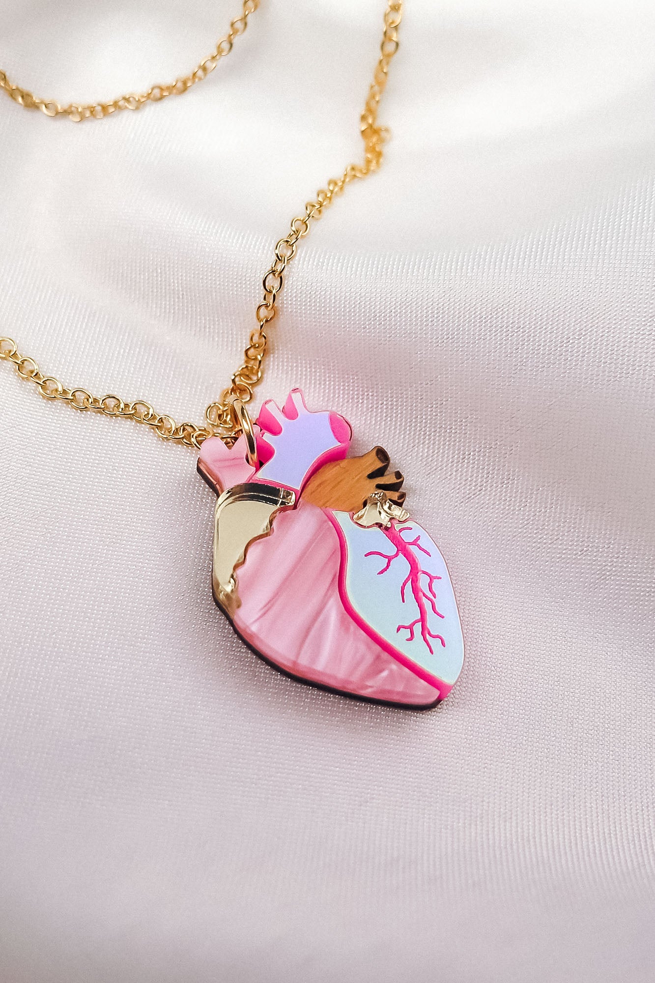 A unique jewelry piece, this gold chain necklace features a pendant shaped like an anatomical heart with pink, white, and gold details, beautifully displayed on soft, light-colored satin fabric.