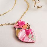 A gold chain anatomical heart necklace with a pendant in pink, mint green, and gold lies on a light surface, surrounded by dried grass—a unique jewelry piece for lovers of handcrafted jewelry.