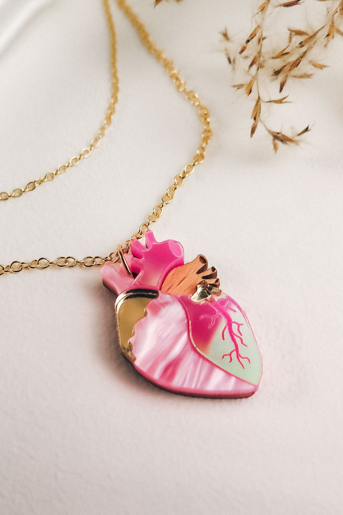 A gold chain anatomical heart necklace with a pendant in pink, mint green, and gold lies on a light surface, surrounded by dried grass—a unique jewelry piece for lovers of handcrafted jewelry.