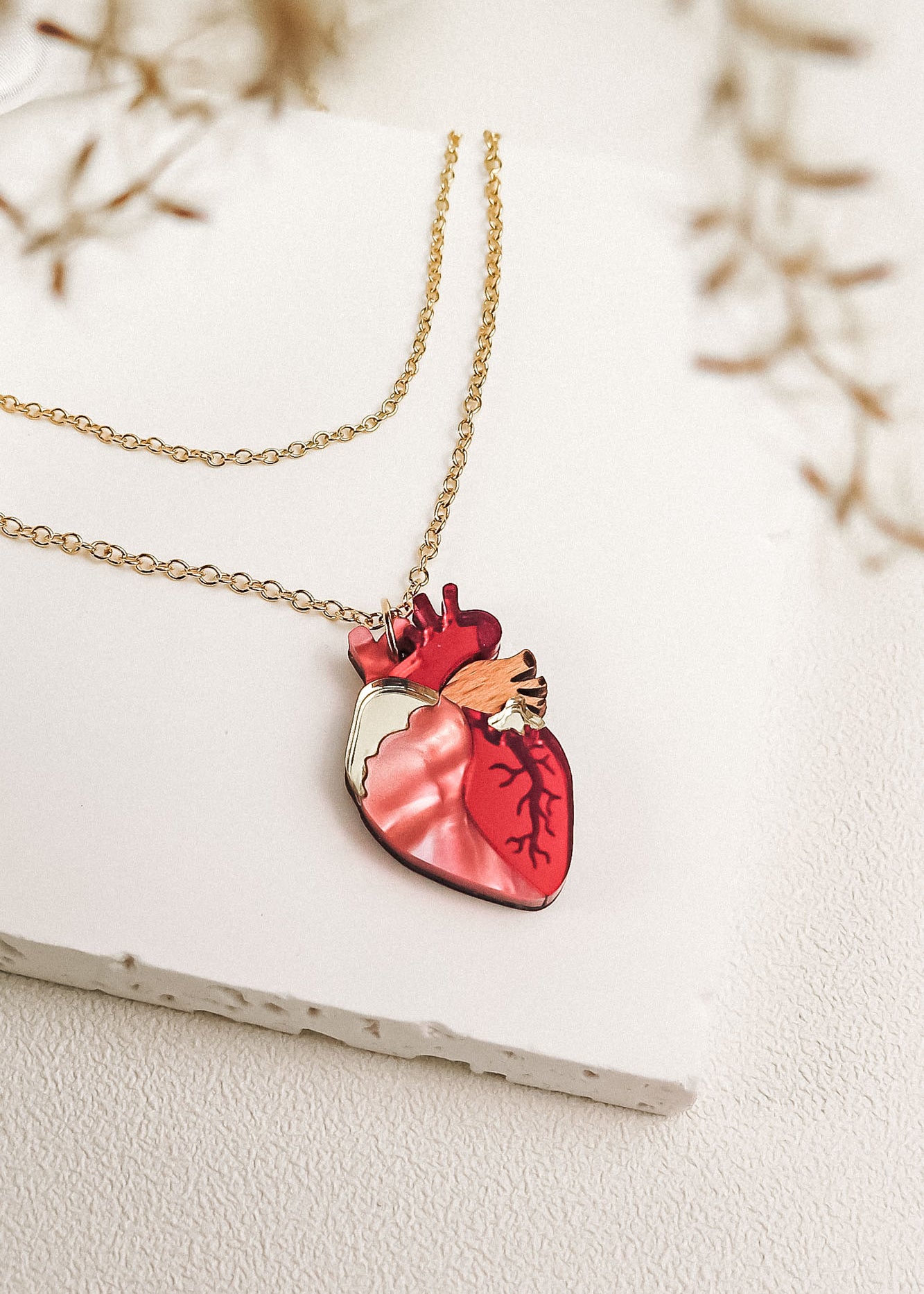 A gold chain anatomical heart necklace with a pendant shaped like a human heart, featuring red and pink detailing, is displayed on a white textured surface with delicate blurred foliage in the foreground—a truly unique jewelry piece.
