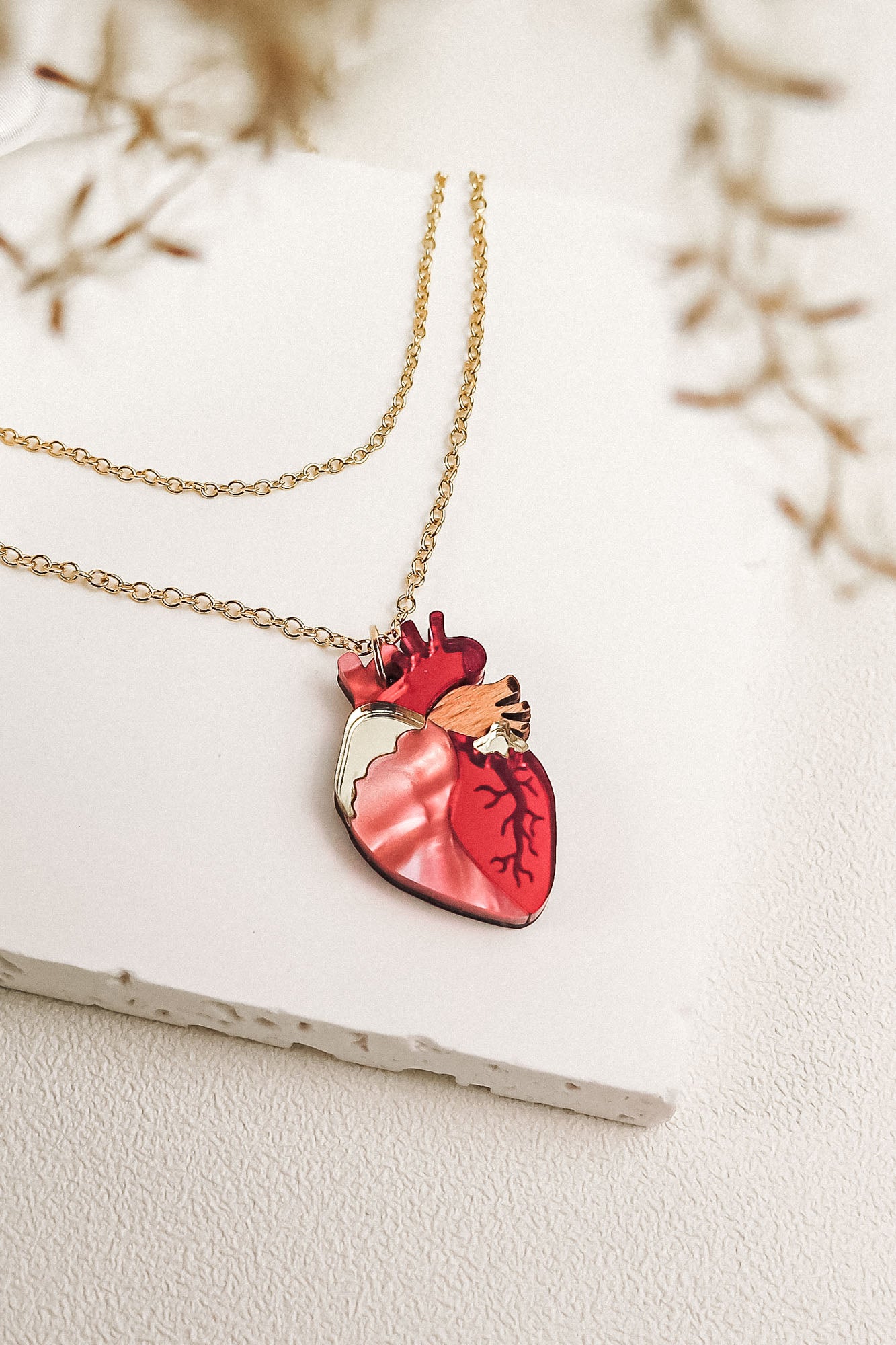 A gold chain anatomical heart necklace with a pendant shaped like a human heart, featuring red and pink detailing, is displayed on a white textured surface with delicate blurred foliage in the foreground—a truly unique jewelry piece.