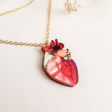 A gold chain anatomical heart necklace with a pendant in red and pink tones, resting on a light textured surface with dried plant accents—a unique jewelry piece that stands out.