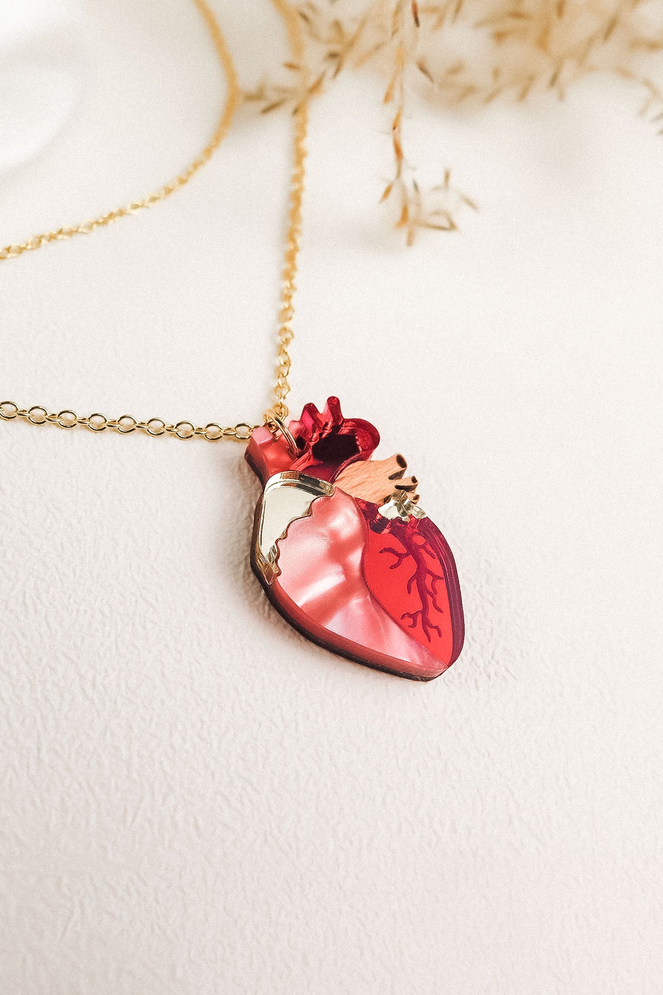 A gold chain anatomical heart necklace with a pendant in red and pink tones, resting on a light textured surface with dried plant accents—a unique jewelry piece that stands out.
