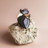 A handcrafted black and gold Anatomical Heart Pin rests on a textured light-colored rock against a plain beige background, making it a true statement piece.