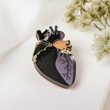 A handcrafted anatomical heart pin with black, purple, gold, and brown details rests on a white surface beside delicate white flowers—a striking statement piece for any collection.